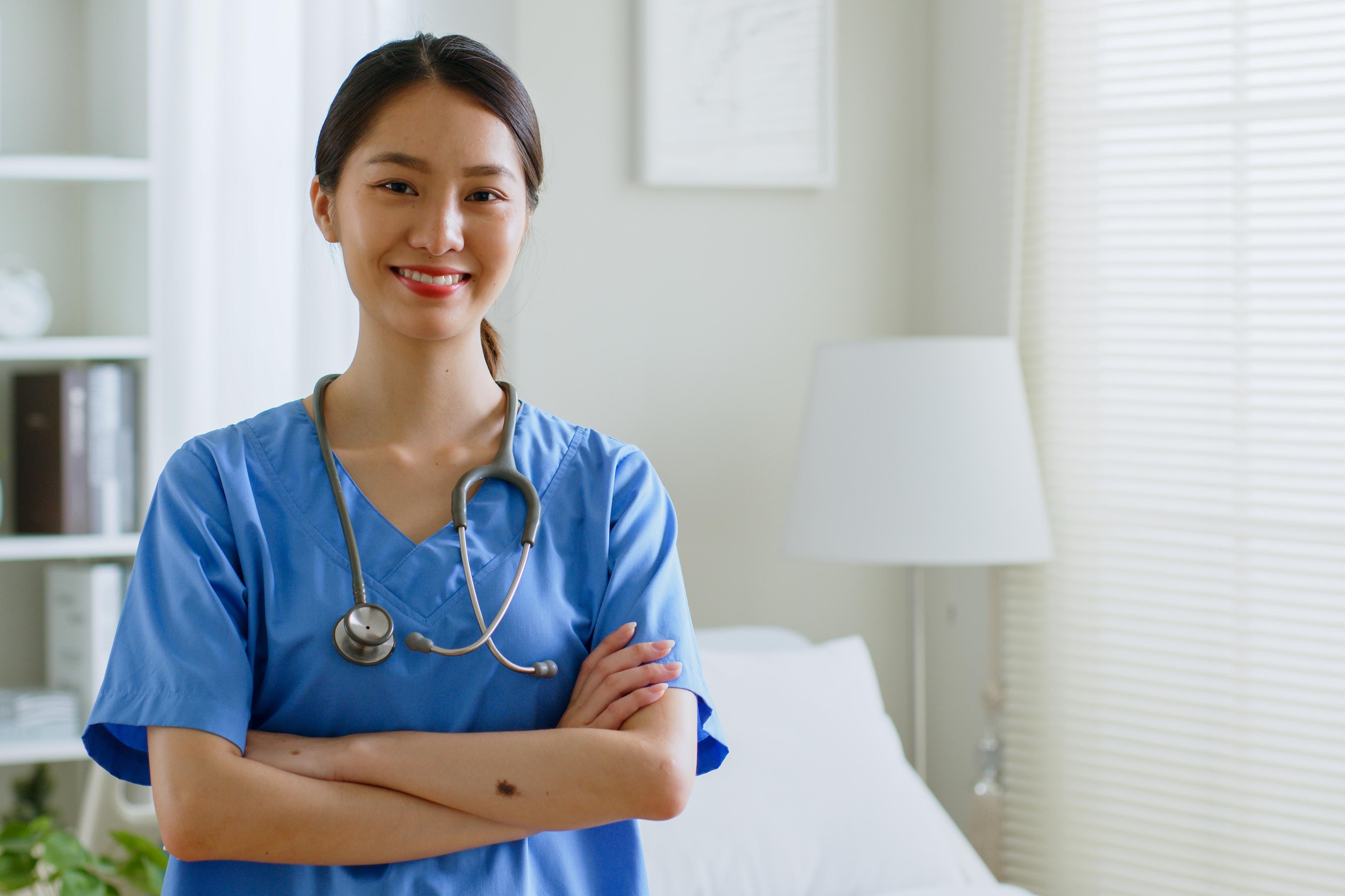Smiling nurse in blue scrubs with a stethoscope around her neck, standing in a bright, tidy room with crossed arms, conveying professionalism and warmth.