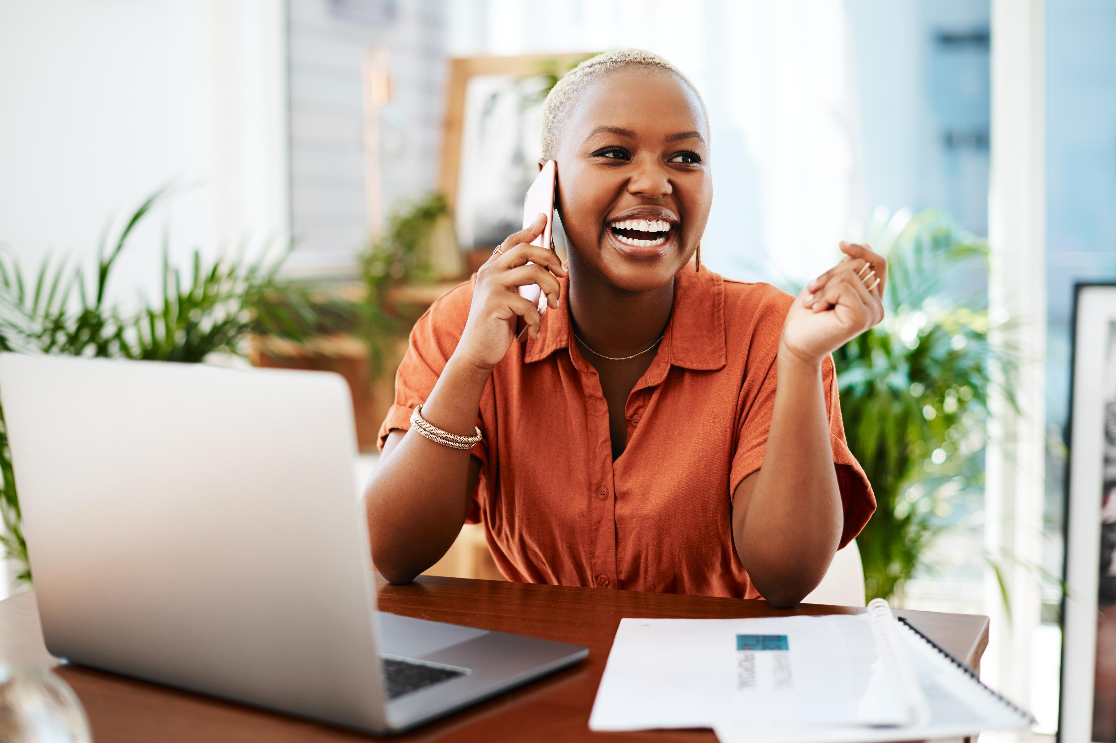 A smiling woman talks on the phone at a desk with a laptop and documents.